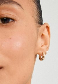 Guld hoop ørering med glat overflade, parret med en lille hvid perle studs. Nærbillede af et øre, der viser begge øreringe.