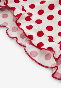 Biela tkanina s červenými bodkami, s volánovým lemom. Volán má ružové akcenty a červený prešitý okraj.