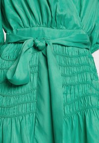 El vestido verde presenta una cintura ceñida con un lazo atado, tejido con textura y un material suave y ligero.