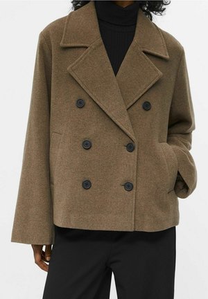 Person wearing a brown double-breasted wool coat over a black turtleneck, with one hand in the coat pocket.