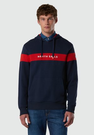 Jeune homme portant un sweat à capuche bleu marine avec une bande rouge et le texte "NORTH SAILS", assorti d'un jean bleu, debout devant un fond uni.