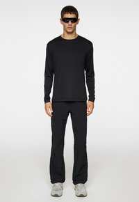 Svart långärmad skjorta och byxor set, tillverkat av lättviktsmaterial. Kombineras med mörka solglasögon och ljusgrå sneakers.