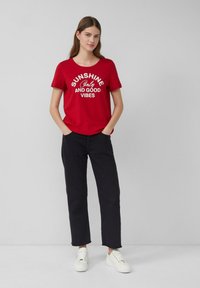 Rode katoenen T-shirt met "SUNSHINE Only AND GOOD VIBES" in wit gedrukt. Gecombineerd met zwarte broeken en witte schoenen. Model kijkt recht vooruit.