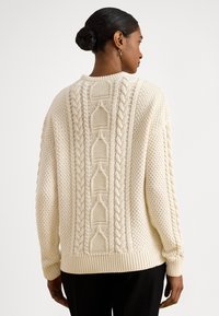 Hrubý krémový pletený sveter s kombinovaným dizajnom textúr, výraznými vzormi kabeliek a rebrovaným lemom, s voľným strihom a spustenými ramenami.