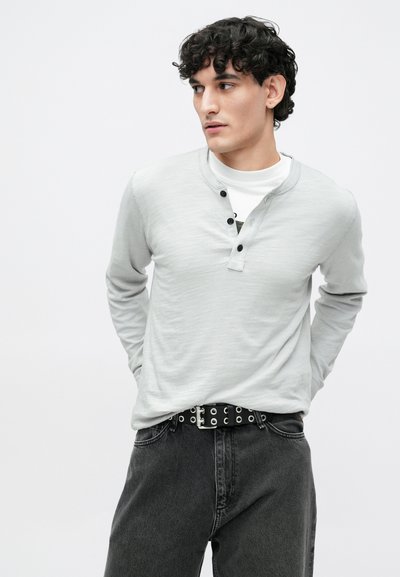 Szara, długorękawowa koszula henley z czarnymi guzikami, w połączeniu z ciemnymi dżinsami i czarnym paskiem z oczkami. Gładka tekstura, casualowy design.