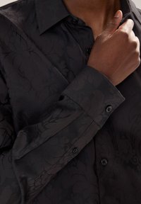 Camicia nera a maniche lunghe con un delicato motivo floreale, caratterizzata da un colletto strutturato, polsini con bottoni e un tessuto liscio e strutturato.