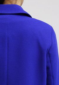 Blazer aderente in un vivace tessuto blu con una finitura testurizzata. Presenta un colletto a rever e linee pulite sulla schiena e sulle maniche.