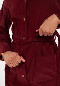 Robe en velours côtelé bourgogne avec un boutonnage à l'avant, une ceinture à la taille et une poche frontale. Présente une texture côtelée et une silhouette décontractée.