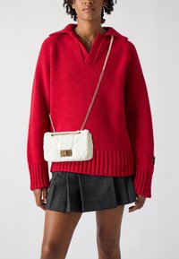 Červený pletený sveter s výstrihom do V a rebrovaným lemom, doplnený bielou prešívanou crossbody taškou s zlatým zámkom a reťazovým popruhom.