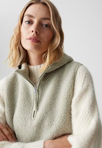 Fleece-Weste in Hellgrün mit hohem Kragen und halbem Reißverschluss. Weiche Textur, kombiniert mit einem cremefarbenen Strickpullover darunter.