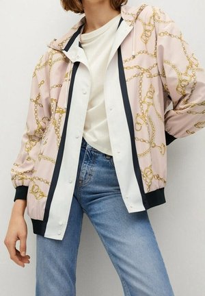 Persoon draagt een lichtroze capuchontrui met goudkettingprint over een wit overhemd met knopen en blauwe spijkerbroek, staand tegen een effen achtergrond.