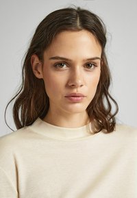 Top beige chiaro con colletto alto, realizzato in un tessuto morbido e testurizzato. Il modello ha capelli ondulati di lunghezza spalla e un trucco naturale.