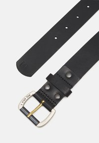 Cintura in pelle nera con fibbia in argento e oro, caratterizzata da borchie e una texture liscia. La punta è affusolata con un logo stampato.