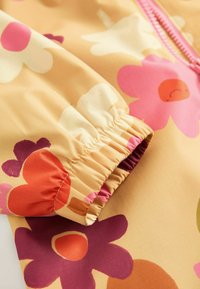 Färgglad regnjacka i blommönster med en beige bakgrund och rosa, röda och lila blommor; samlad elastisk manschettdetalj.