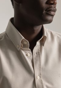 Primer plano de un hombre que lleva una camisa beige abotonada con cuello tipo botón, mostrando parte de su rostro inferior y cuello.