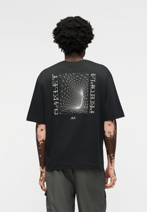 Henkilö, jolla on tummat kiharat hiukset ja vitiligo, pukeutunut mustaan Oakley Prototype -t-paitaan, jossa on valkoinen geometrinen kuvio ja teksti takana.