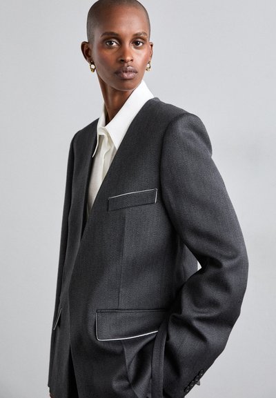 Szürke zakó testreszabott szabással, fehér paszpólós részletekkel, egy köldökzsebbel és textúrált anyagból készült. Fehér ing fölé viselve.