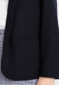 Tmavě modrý blazer s hladkým povrchem, který má jedno kapsu na hrudi a jednoduchý design klopy. Částečně viditelný kostkovaný vzor dole.
