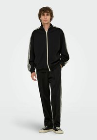 Schwarzer Trainingsanzug mit einer Jacke mit Reißverschluss und Hose, die weiße Seitenstreifen aufweist. Hergestellt aus einem glatten, leichten Material. Lässige Passform.