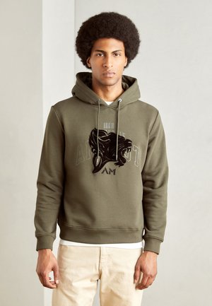 Hoodie vert olive avec un graphique frontal d'une tête de lion et un texte. Possède une capuche avec cordon de serrage et des poignets côtelés. Porté par-dessus une chemise blanche.