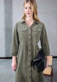 Abito camicia a maniche lunghe verde oliva con apertura a bottoni, due tasche sul petto e vita arricciata, abbinato a una borsa nera oversize.