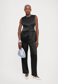 Sieviete, tērpusies melnā bez piedurknēm pogājamo žaketi un atbilstošos melnos biksēs, tur gaismas zilo somu un valkā baltas norādītās papēžkurpes.