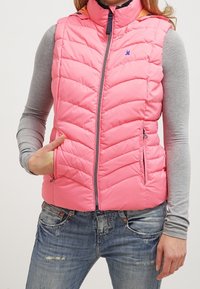 Rosa pufferväst med hög krage, horisontell sömnad, framzipper och två sidofickor. Bärs över en långärmad grå tröja.