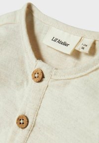 Bež pamukasta košulja s okruglim izrezom, koja ima drveni gumb za zatvaranje i oznaku "Lil' Atelier." Oznaka veličine: 1-2M.
