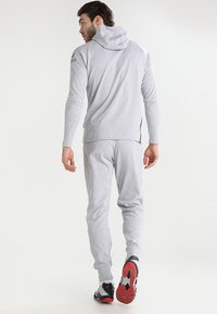 Helles graues Sportanzug mit Kapuze und langen Ärmeln, ausgestattet mit einer glatten Textur, seitlichen Reißverschlüssen und schwarzen Akzenten an den Schuhen.
