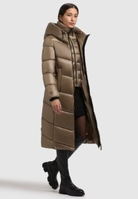 Long manteau en duvet de couleur bronze métallique avec une capuche, design matelassé, fermeture éclair à l'avant et accents noirs. Porté avec des bottes noires jusqu'aux genoux.