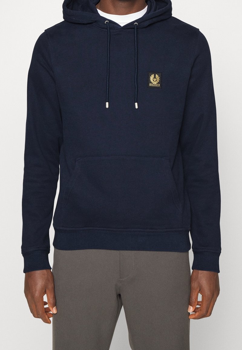 Homme portant un sweat à capuche bleu marine avec poche avant et cordons, associé à un pantalon gris sur un fond uni.