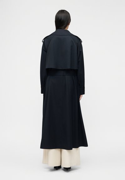 Femme debout, de dos, portant un long trench-coat noir sur un pantalon large beige et des chaussures noires à talons, sur un fond blanc uni.