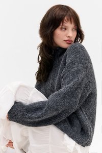 Sweater de malha oversized em cinza escuro com gola alta e fio texturizado. O modelo está sentado com uma peça branca sobreposta, criando contraste.