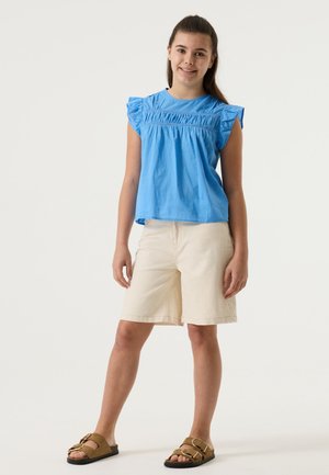 Lichtblauw shirt met korte mouwen en geplooide details, gecombineerd met beige korte broek en bruine sandalen met dubbele gespsluitingen.