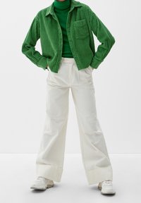 Grüne Kordjacke mit Brusttasche, über einem grünen Rollkragenpullover getragen. Weiße weite Hose, kombiniert mit weißen Sneakers.