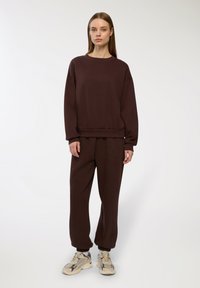 Brązowa bluza z kapturem i dopasowane dresy w luźnym kroju, wykonane z miękkiego materiału. Okrągły dekolt, elastyczne mankiety i pasek w talii. Sneakersy z jasnymi akcentami.