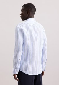 Camicia a maniche lunghe in cotone leggero con righe verticali blu e bianche. Presenta un colletto puntato e polsini con bottoni. L'orlo è leggermente curvato.