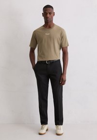 Man staat tegen een effen muur, gekleed in een olijfgroene t-shirt, zwarte broek met een riem, en witte sneakers met gumkleurige zolen.