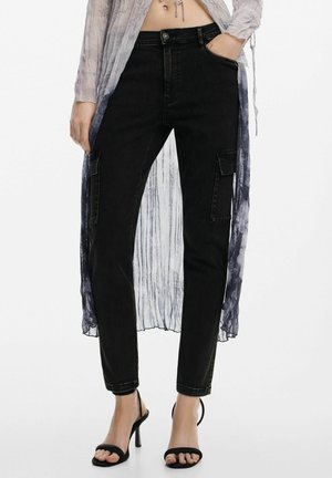 Frau trägt schwarze, hoch taillierte Cargo-Hosen mit Klappentaschen, transparente, langärmelige Ombre-Bluse und schwarze Sandalen mit Absatz, steht mit einer Hand in der Tasche.