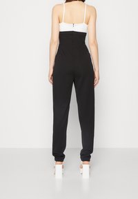 Zwarte jumpsuit met nauwsluitend bovenstuk, witte bandjes en taps toelopende pijpen. Voorzien van een rits aan de achterkant en een gladde stof met een matte afwerking.