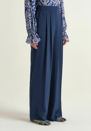 Personne portant un pantalon large taille haute bleu marine associé à un chemisier bleu à manches longues à motifs et des chaussures plates multicolores.