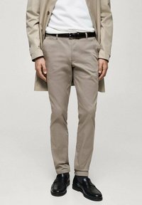 Světlé béžové kalhoty z měkké látky, s přizpůsobeným střihem a hladkou texturou. Nošeny s bílým svetrem a černými koženými botami.