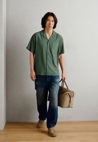 Green striped short-sleeve shirt, blue jeans, beige bag, tan shoes, standing on wooden floor with gray wall backdrop.