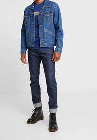 Blå jeansjacka med två bröstfickor och kontrasterande sömmar, kombinerad med mörka jeans och svarta snörstövlar med uppvikta muddar.