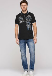 Czarny polo z białymi i niebieskimi akcentami, z graficznym wzorem tekstowym. W połączeniu z jasnoniebieskimi dżinsami i białymi sneakersami.