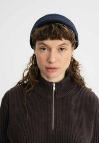 Donkerbruine geribde trui met een hoge kraag en een zilveren rits. Het model draagt een marineblauwe gebreide muts met textuurpatroon details.