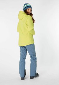 Heldere gele waterdichte jas met capuchon en zijritsen, gecombineerd met blauwe waterdichte broek. Gebreide blauwe beanie met groene strepen.