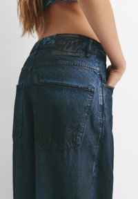 Džínsy z denimu v tmavomodrej farbe s vyblednutou textúrou. Majú čiernu nášivku na páse a dva veľké zadné vrecká s prešitým detailom.