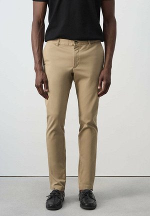 Mann trägt beige, schlanke Hose, schwarzes Hemd und schwarze Schnürschuhe, steht vor einem einfarbigen hellen Hintergrund.