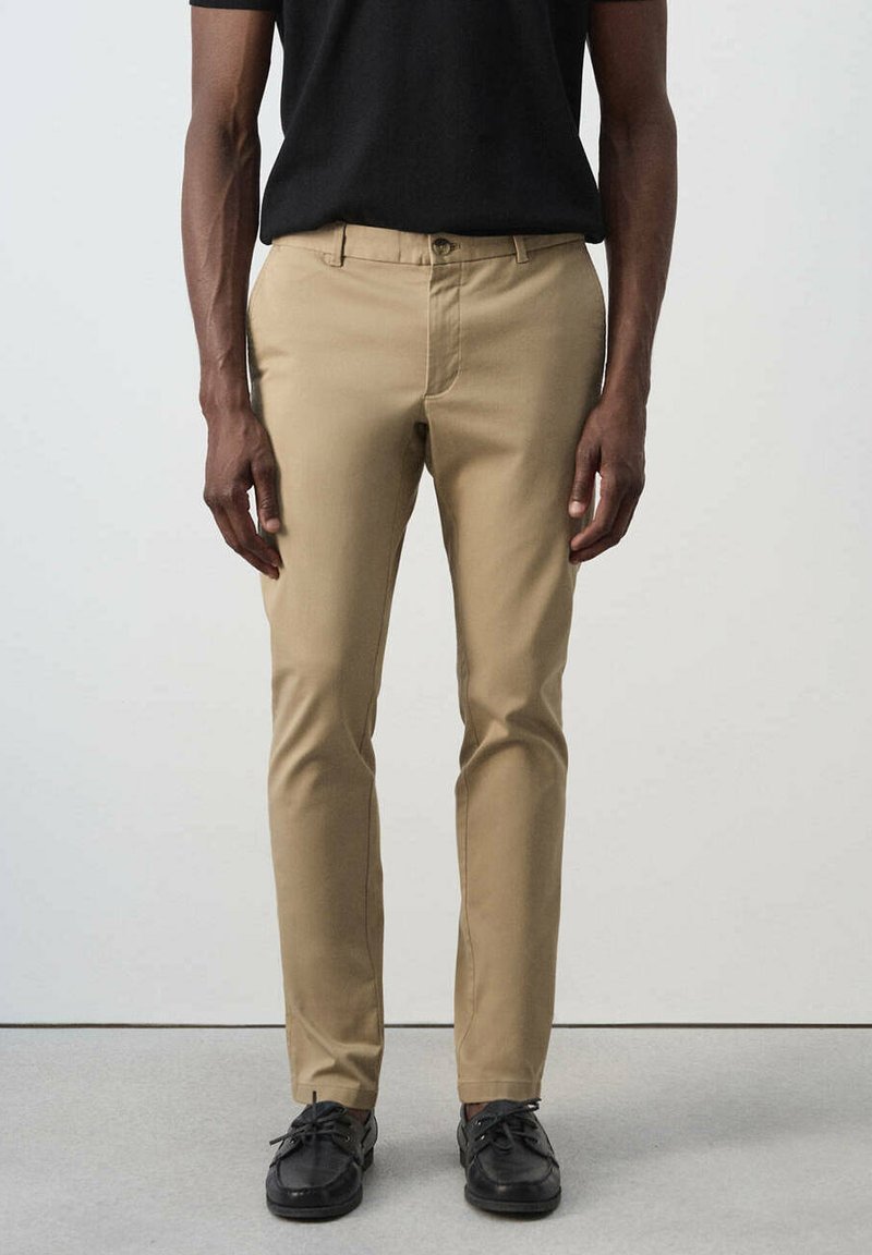 Homme portant un pantalon beige slim, une chemise noire et des chaussures noires à lacets, debout devant un fond clair uni.
